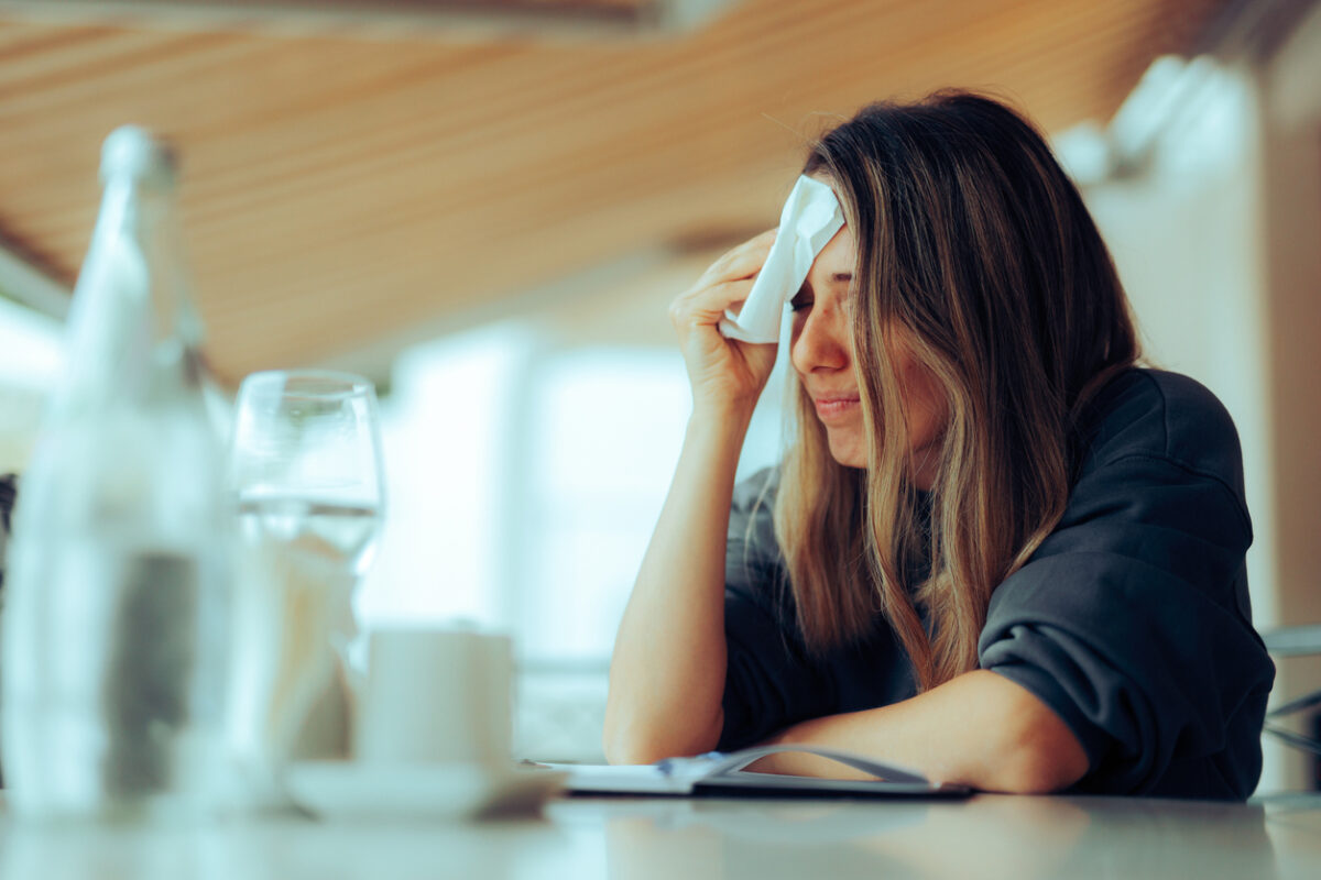 Woman Wiping Sweaty Forehead Using a Napkin. She sits at a table set with water glasses.