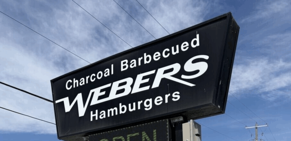 A black sign with white lettering reading Webers hamburgers. Behind is a blue sky with wispy clouds