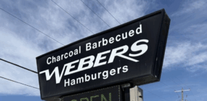 A black sign with white lettering reading Webers hamburgers. Behind is a blue sky with wispy clouds